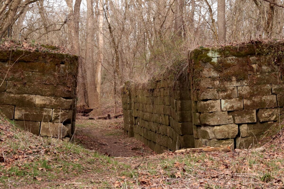 Stone ruins of a canal lock stand in a wooded area, with large stone blocks forming two parallel walls, and a dirt path running between them. Leafless trees can be seen in the background, and dry leaves cover the ground.